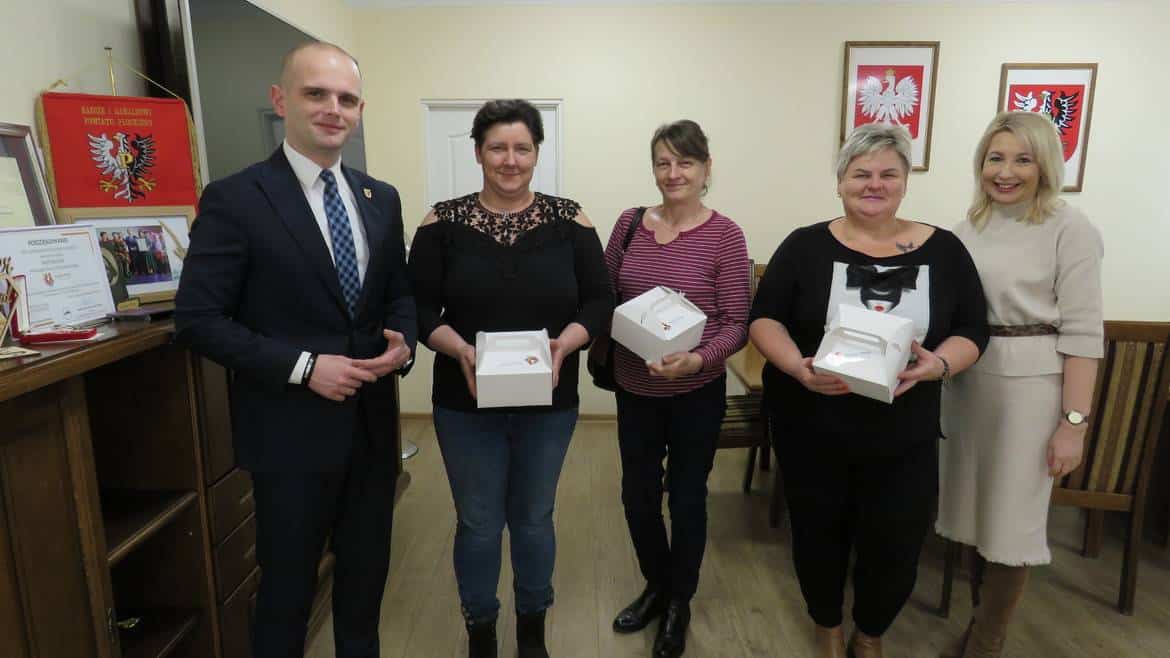 Na zdjęciu widać grupę pięciu osób, w tym mężczyznę w garniturze oraz cztery kobiety, które trzymają białe pudełka, w tle widoczne są flagi i dyplomy.