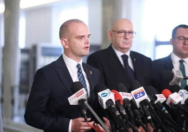 Na zdjęciu widać mężczyznę w garniturze, przemawiającego do dziennikarzy przed mikrofonami, z dwoma innymi mężczyznami stojącymi obok.