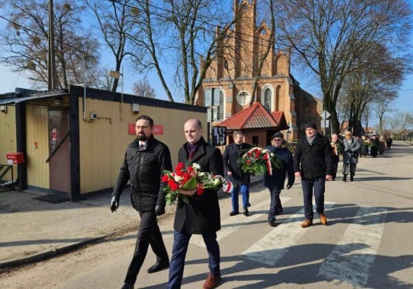 Grupa mężczyzn w garniturach i płaszczach niesie kwiaty w kierunku kościoła podczas uroczystości.