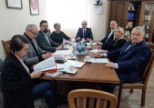 Grupa ludzi siedzi przy stole konferencyjnym, uczestnicząc w spotkaniu, z dokumentami i filiżankami na stole.