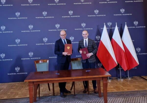 Dwaj mężczyźni w garniturach trzymają dokumenty, stojąc przy stole w sali podpisania umowy, z flagami Polski w tle.