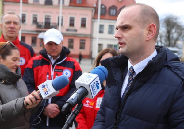 Na zdjęciu widoczny jest mężczyzna w ciemnym płaszczu, przemawiający do mikrofonu, w towarzystwie osób ubranych w czerwone kurtki, podczas wydarzenia publicznego na świeżym powietrzu.