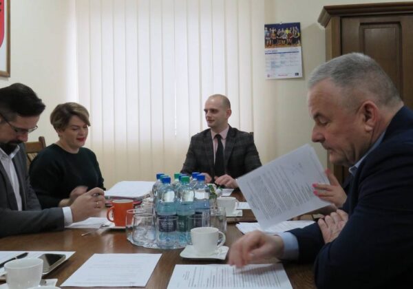 Na zdjęciu widać grupę osób zebranych przy stole, prowadzących dyskusję w biurze, z dokumentami i napojami na stole.