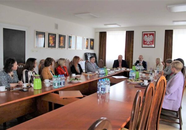 Spotkanie w sali konferencyjnej z uczestnikami siedzącymi przy długim stole, w otoczeniu obrazów i z podaną kawą oraz jedzeniem.