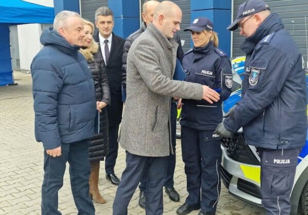 Na zdjęciu widoczna jest grupa osób w strojach cywilnych oraz mundurach policyjnych, którzy rozmawiają przy pojeździe policyjnym.