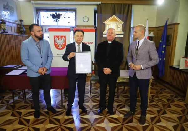 Na zdjęciu czterech mężczyzn stoi w sali, trzymając dyplom, podczas ceremonii wręczenia nagrody.