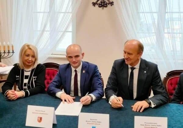 Na zdjęciu czterech mężczyzn i jedna kobieta uczestniczą w ceremonii podpisania dokumentów w eleganckim wnętrzu.