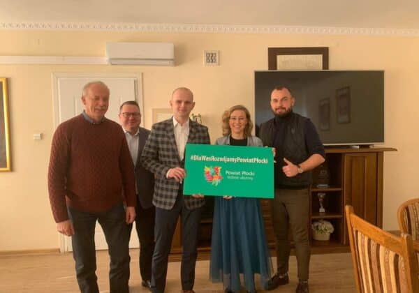 Grupa ludzi trzymająca zielony plakat promujący rozwój Powiatu Płockiego, pozując na tle eleganckiego wnętrza.