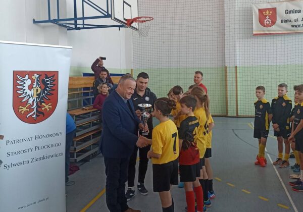 Na zdjęciu widoczna jest ceremonia wręczenia pucharu dzieciom w koszulkach sportowych, odbywająca się w hali sportowej, z przedstawicielami władz lokalnych.