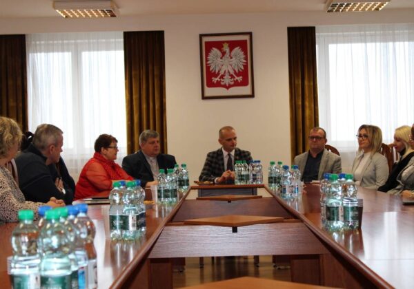 Na zdjęciu przedstawiono spotkanie grupy ludzi siedzących wokół stołu w sali konferencyjnej, z herbem Polski na ścianie w tle.
