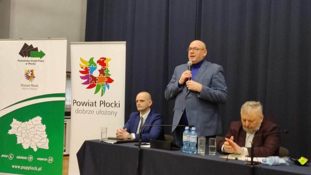 Na zdjęciu widoczni są mężczyźni podczas wystąpienia na spotkaniu związanym z Powiatowym Urzędem Pracy w Płocku, stojąc obok bannerów promujących powiat.