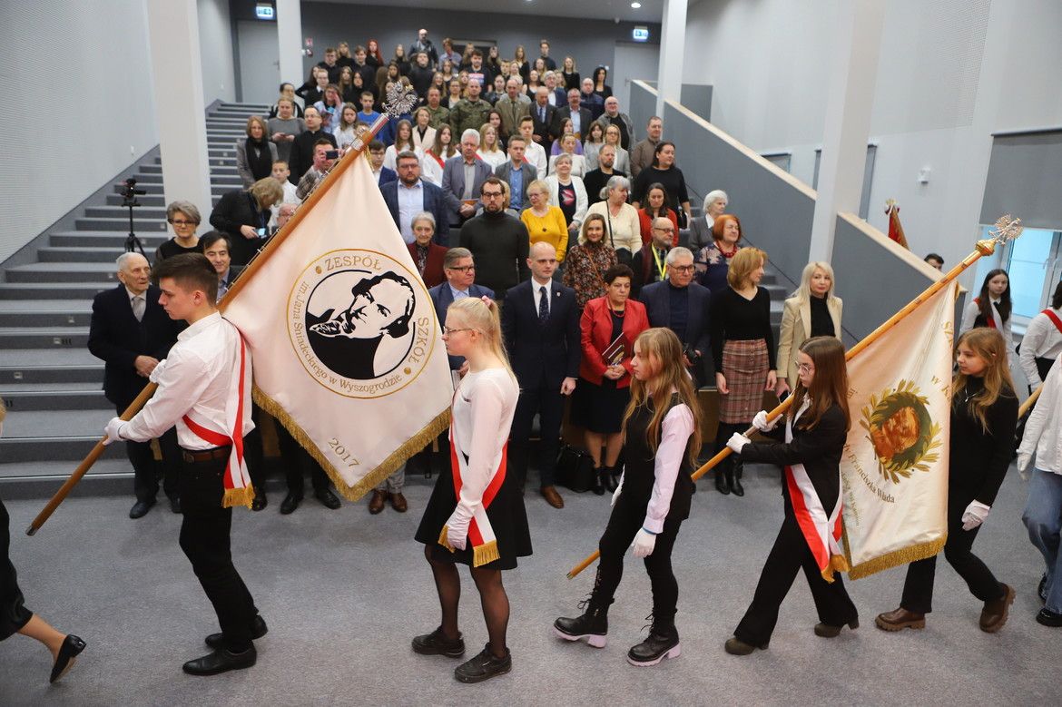 Uroczystość szkolna z pocztem sztandarowym i zgromadzonymi gośćmi w nowoczesnej auli.
