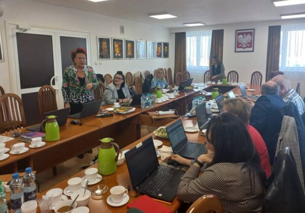 Na zdjęciu widać spotkanie robocze w sali konferencyjnej, gdzie uczestnicy pracują na laptopach, a jedna osoba prowadzi prezentację.