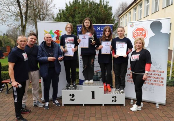 Na zdjęciu znajdują się zwycięzcy konkursu sportowego, stojący na podium z dyplomami i trofeami, we wspólnym ujęciu z organizatorami wydarzenia.
