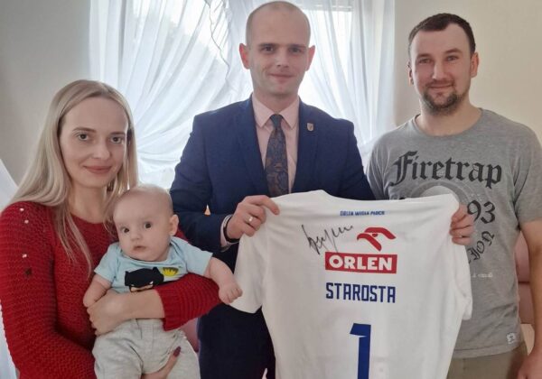 Na zdjęciu znajduje się rodzina pozująca z podpisanym koszulą sportową, w której matka trzyma niemowlę, a obok niej mężczyzna w garniturze oraz mężczyzna w szarej koszulce.