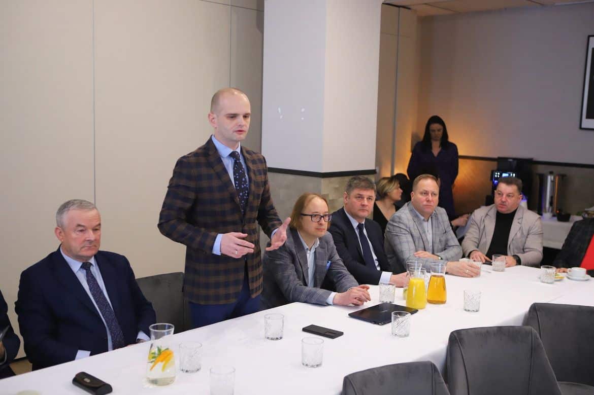 W trakcie spotkania biznesowego mężczyzna w garniturze przemawia do zgromadzonych przy stole, wśród których siedzą inni elegancko ubrani uczestnicy.