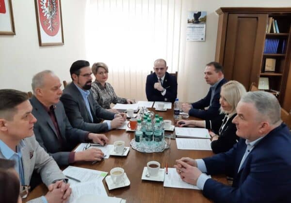 Na zdjęciu przedstawiono zebranie grupy osób z różnymi dokumentami i napojami, w przeszklonym pomieszczeniu z widocznymi herbami oraz kalendarzem.