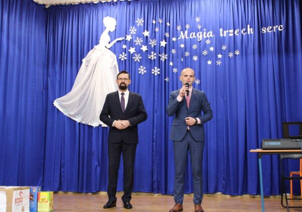 Na zdjęciu dwóch mężczyzn w eleganckich garniturach stoi na scenie przy niebieskim tle z dekoracjami przedstawiającymi postać kobiety oraz napisem "Magia trzech serc".