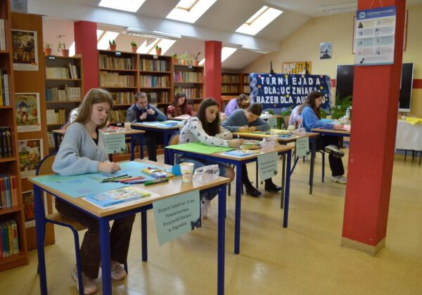 Uczestnicy turnieju plastycznego pracują nad swoimi projektami w bibliotece, otoczeni książkami i materiałami artystycznymi.