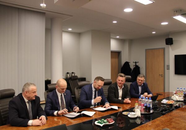 Na zdjęciu pięciu mężczyzn siedzi przy stole w sali konferencyjnej, podpisując dokumenty, przy czym na stole znajdują się napoje i przekąski.