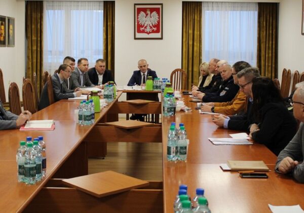 Na zdjęciu przedstawiono posiedzenie w sali konferencyjnej, podczas którego grupa osób dyskutuje przy długim stole.