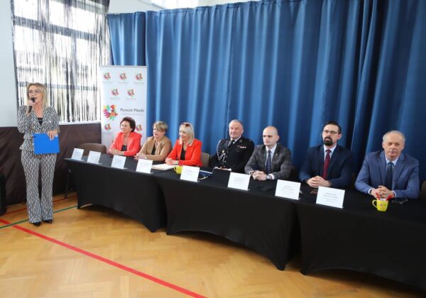 Na zdjęciu widać grupę osób siedzących przy stole, z jedną kobietą mówiącą do mikrofonu, podczas wydarzenia organizowanego przez Powiat Płocki.