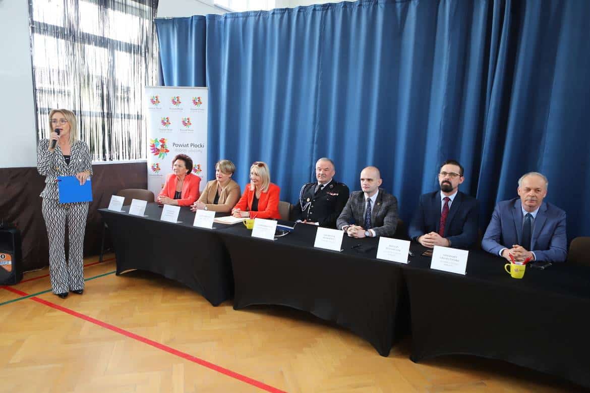 Na zdjęciu widać grupę osób siedzących przy stole, z jedną kobietą mówiącą do mikrofonu, podczas wydarzenia organizowanego przez Powiat Płocki.