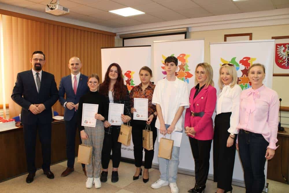 Na zdjęciu grupa osób otrzymuje podziękowania za swoją działalność, stojąc w sali z certyfikatami i torebkami, w otoczeniu przedstawicieli lokalnych władz.