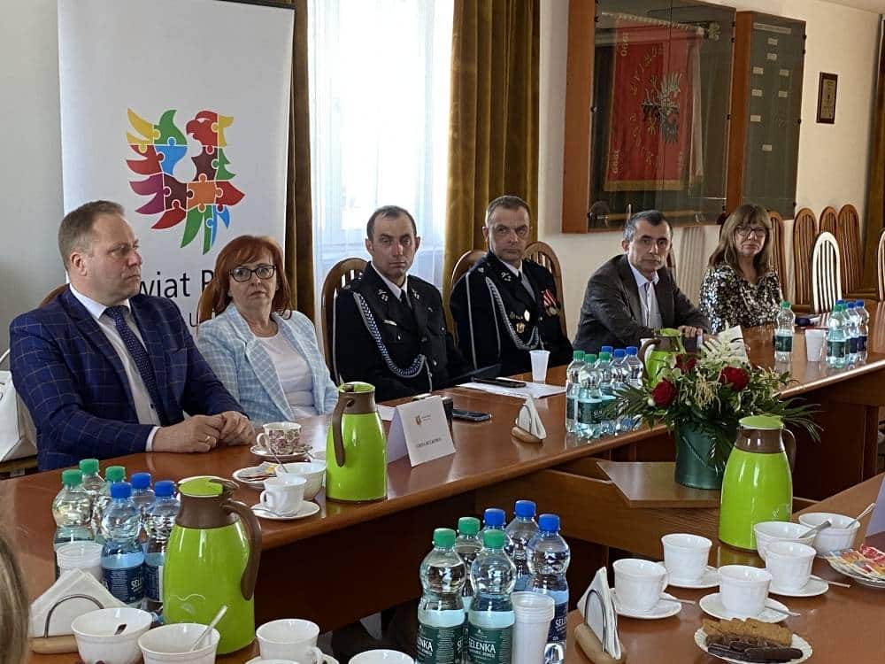 Na zdjęciu widoczna jest grupa ludzi siedzących przy stole, podczas wydarzenia związanego z wsparciem dla Ochotniczej Straży Pożarnej, z logo Powiatu Płockiego w tle.