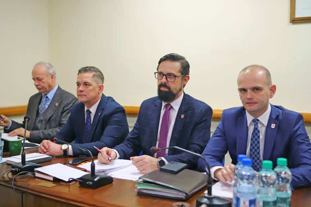 Czterech mężczyzn siedzących przy stole podczas sesji Rady Powiatu, z notesami i mikrofonami przed sobą.