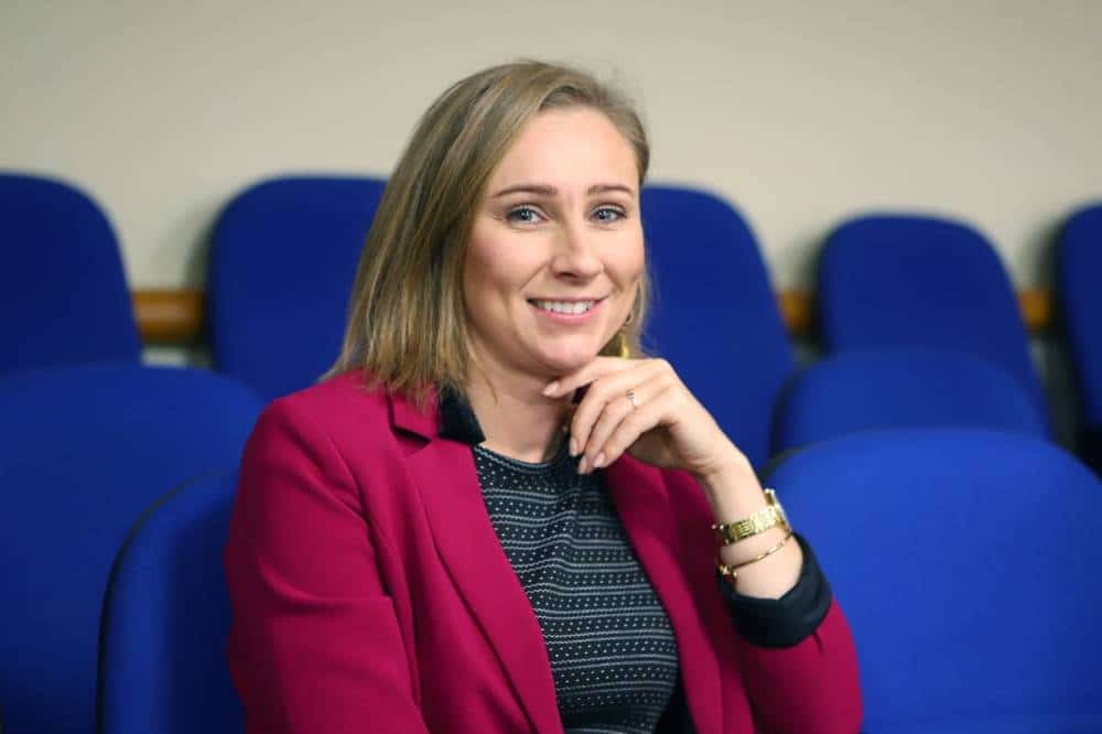 Na zdjęciu widoczna jest uśmiechnięta kobieta w różowej marynarce, siedząca na tle niebieskich krzeseł.