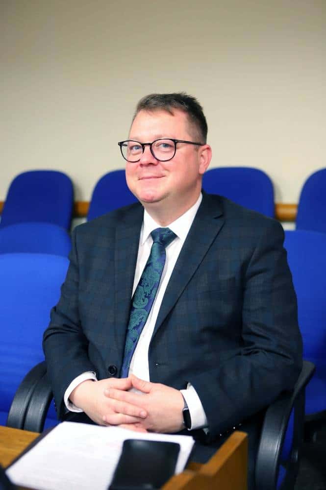 Mężczyzna w okularach siedzi na sali obrad, ubrany w garnitur i krawat, z uśmiechem na twarzy.