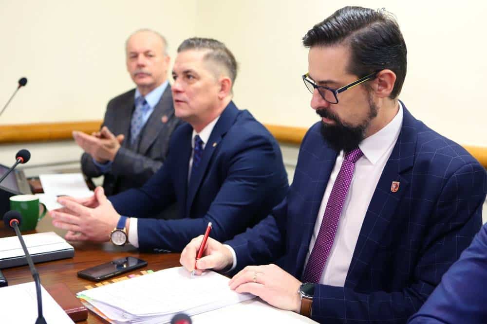 Na zdjęciu widać trzech mężczyzn siedzących przy stole, którzy uczestniczą w sesji rady powiatu, a jeden z nich skupiony jest na pisaniu.