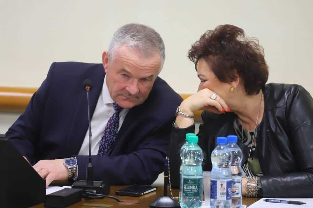 Dwoje ludzi siedzi przy stole, rozmawiając i uśmiechając się do siebie podczas sesji rady powiatu.