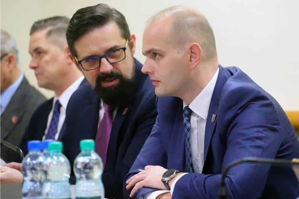 Na zdjęciu widoczni są mężczyźni w formalnych garniturach, z zaangażowanymi wyrazami twarzy, rozmawiający na spotkaniu.