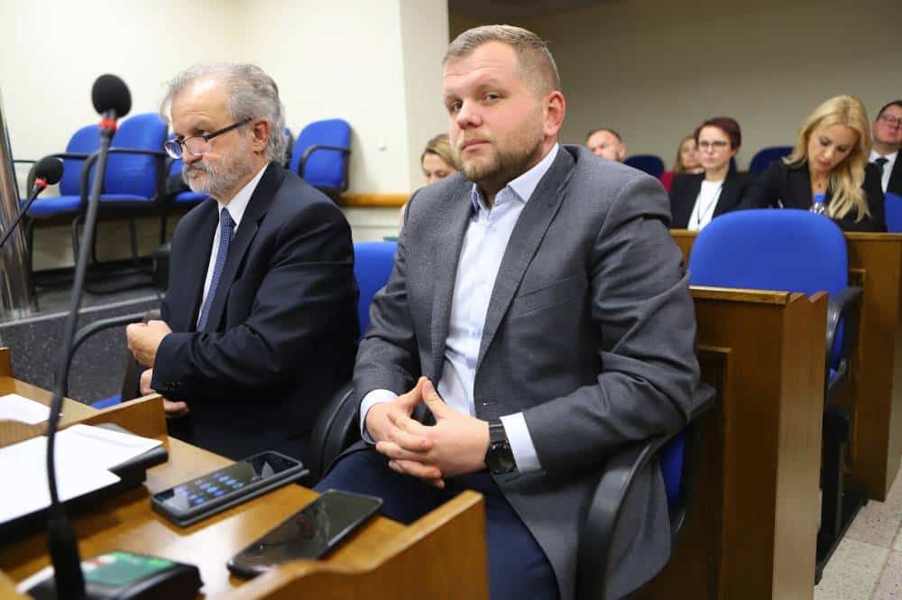 Na zdjęciu widoczna jest dwójka mężczyzn siedzących przy stole w sali obrad, z mikrofonami i sprzętem nagłaśniającym w tle.