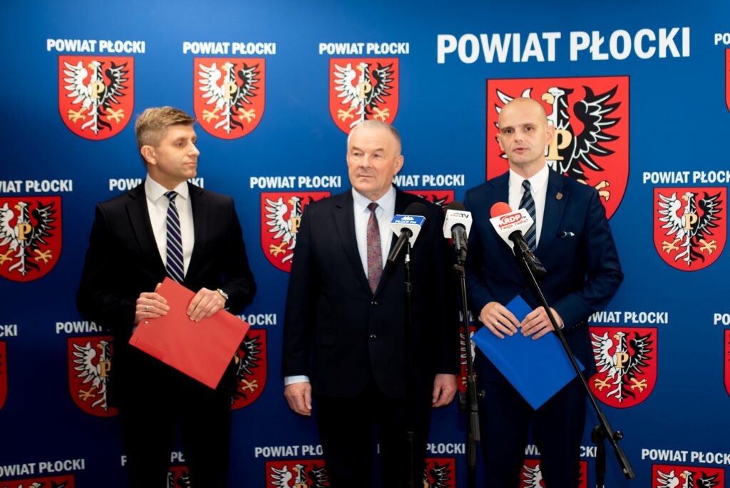 Na zdjęciu trzej mężczyźni w garniturach stoją przed logo Powiatu Płockiego, trzymając dokumenty, podczas konferencji o nowych inwestycjach związanych z partnerstwem samorządu z Bankiem Gospodarstwa Krajowego.