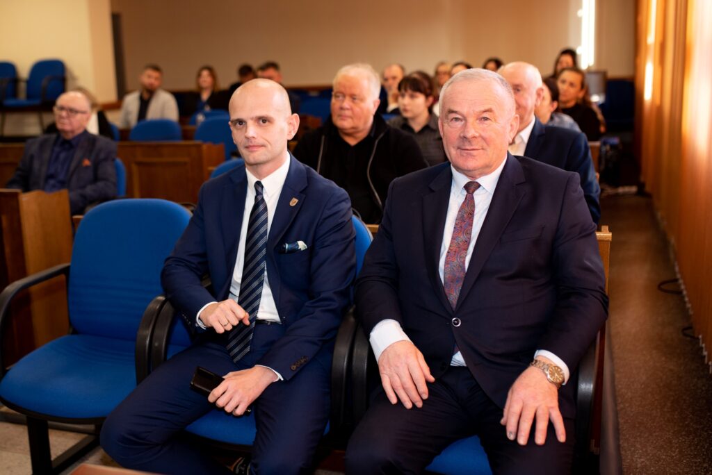 Na zdjęciu widać dwóch mężczyzn w garniturach siedzących w sali, otoczonych innymi uczestnikami spotkania dotyczącego nowych inwestycji i współpracy samorządu z Bankiem Gospodarstwa Krajowego.