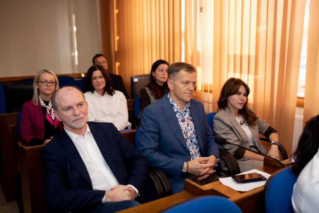 Na zdjęciu widać grupę ludzi siedzących w sali konferencyjnej, podczas spotkania dotyczącego nowych inwestycji samorządowych.