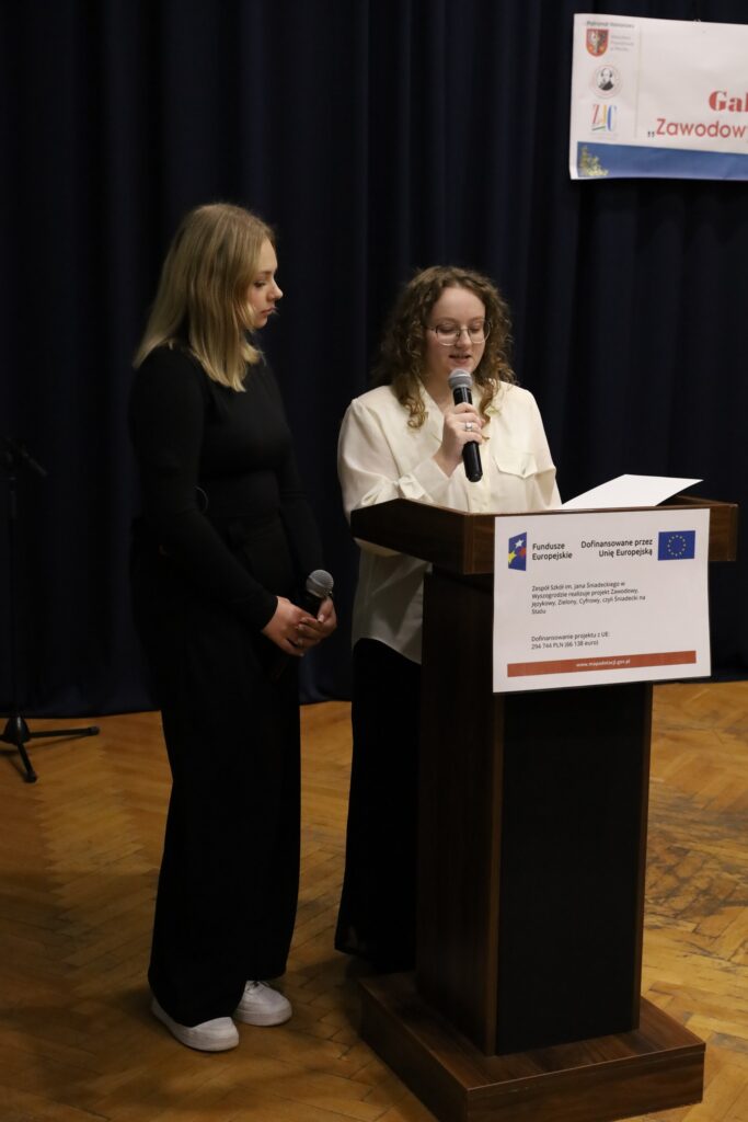 Na zdjęciu dwie młode kobiety, jedna z mikrofonem, a druga obok niej, stoją przy mównicy na gali podsumowującej projekt "Zawodowy, Językowy, Zielony, Cyfrowy, czyli Śniadecki na Stażu".