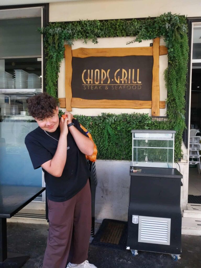 Na zdjęciu widać młodego chłopca pozującego przed restauracją "CHOPS.GRILL", w tle z widocznym zielonym wystrojem oraz logo lokalu.