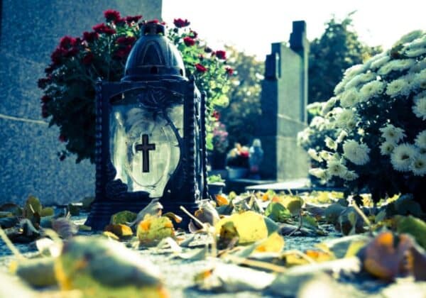 Na zdjęciu widać znicz z krzyżem na cmentarzu, otoczony opadłymi liśćmi i kwiatami.