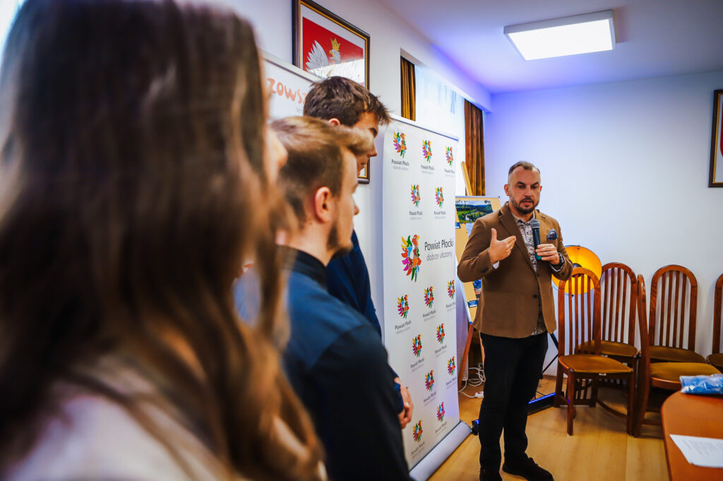 Na zdjęciu widać młodzieżowych radnych oraz osobę przemawiającą podczas ceremonii wręczenia nagród laureatom konkursu organizowanego przez Powiat Płocki.