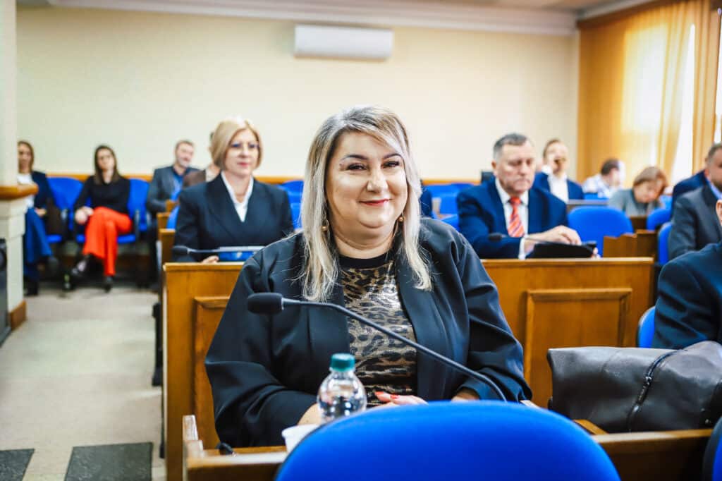 Na zdjęciu przedstawiono uczestników XVI Sesji Rady Powiatu w Płocku, z uśmiechniętą kobietą w pierwszym planie, otoczoną innymi członkami rady.