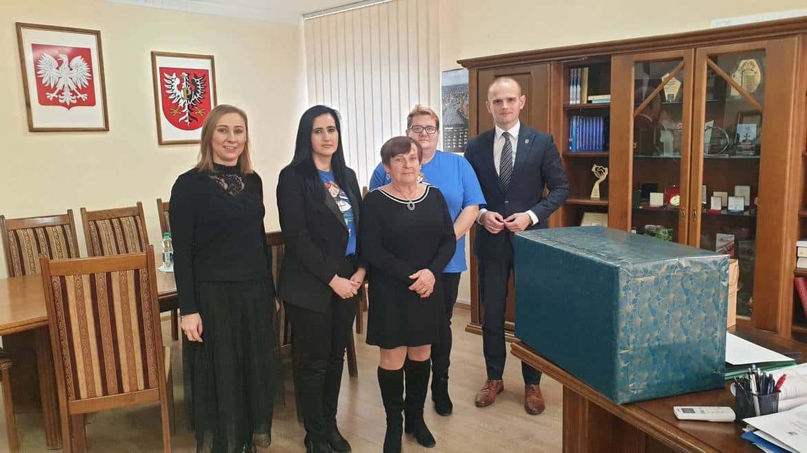 Na zdjęciu przedstawiono pięć osób stojących obok siebie w biurze, z emblematami Polski na ścianie oraz z zawiniątkiem w tle.