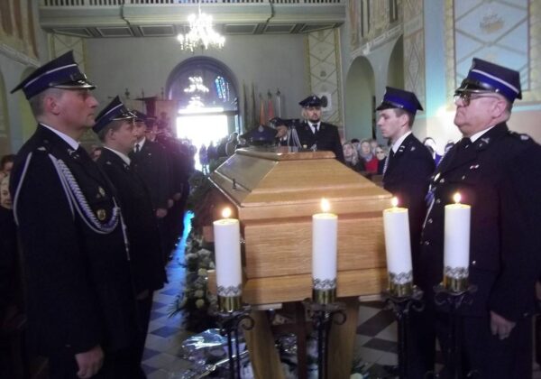 Na zdjęciu widoczny jest pogrzeb z udziałem strażaków, którzy stoją w konsternacyjnej postawie przed trumną, przy której palą się świece.