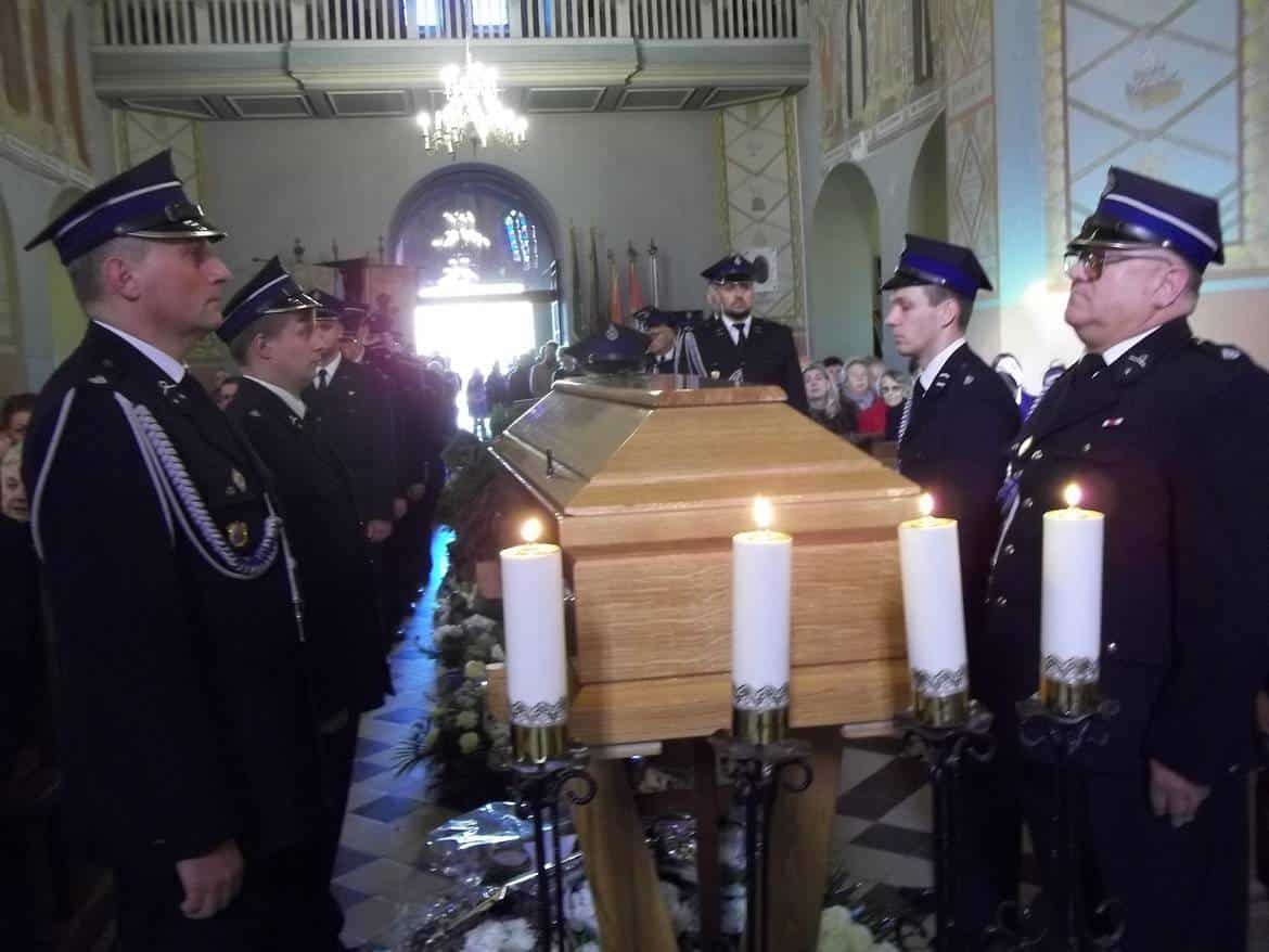 Na zdjęciu widoczny jest pogrzeb z udziałem strażaków, którzy stoją w konsternacyjnej postawie przed trumną, przy której palą się świece.