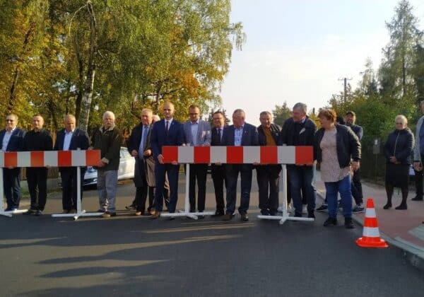 Na zdjęciu widoczna jest grupa osób stojących przy barierkach na drodze, w tle drzewa w jesiennych kolorach.