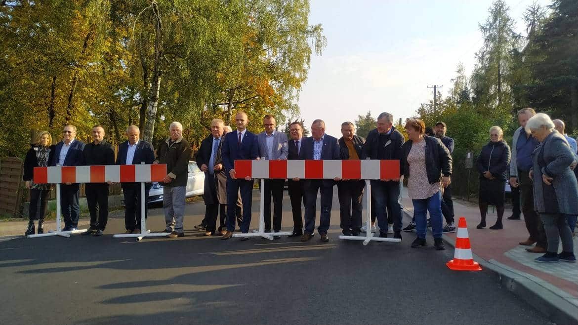 Na zdjęciu widoczna jest grupa osób stojących przy barierkach na drodze, w tle drzewa w jesiennych kolorach.