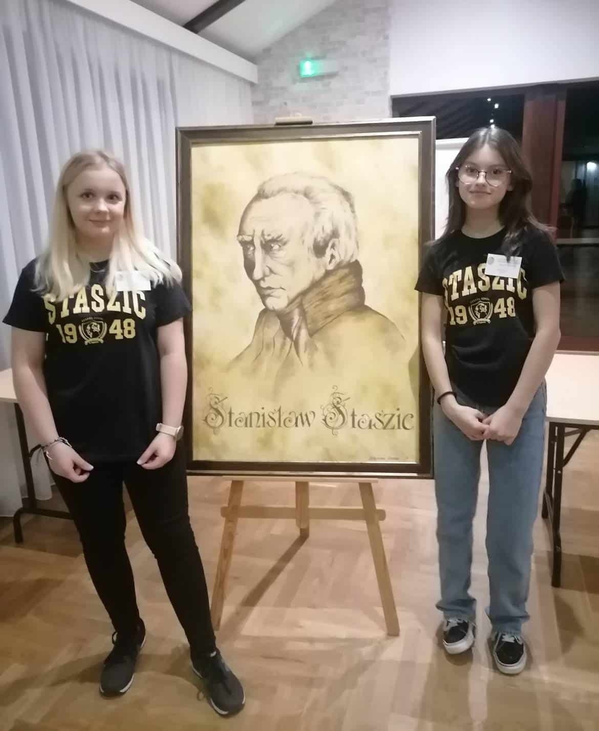 Na zdjęciu dwie młode kobiety stoją obok portretu Stanisława Staszica.